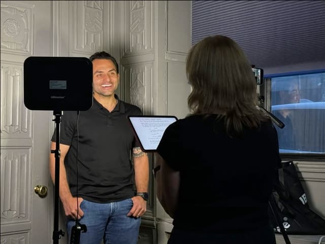 Cathy Reinking, seen from behind with a script on a tablet, gives a redirect to actor Chris Anthony Hernandez, who stands on camera under a light panel.