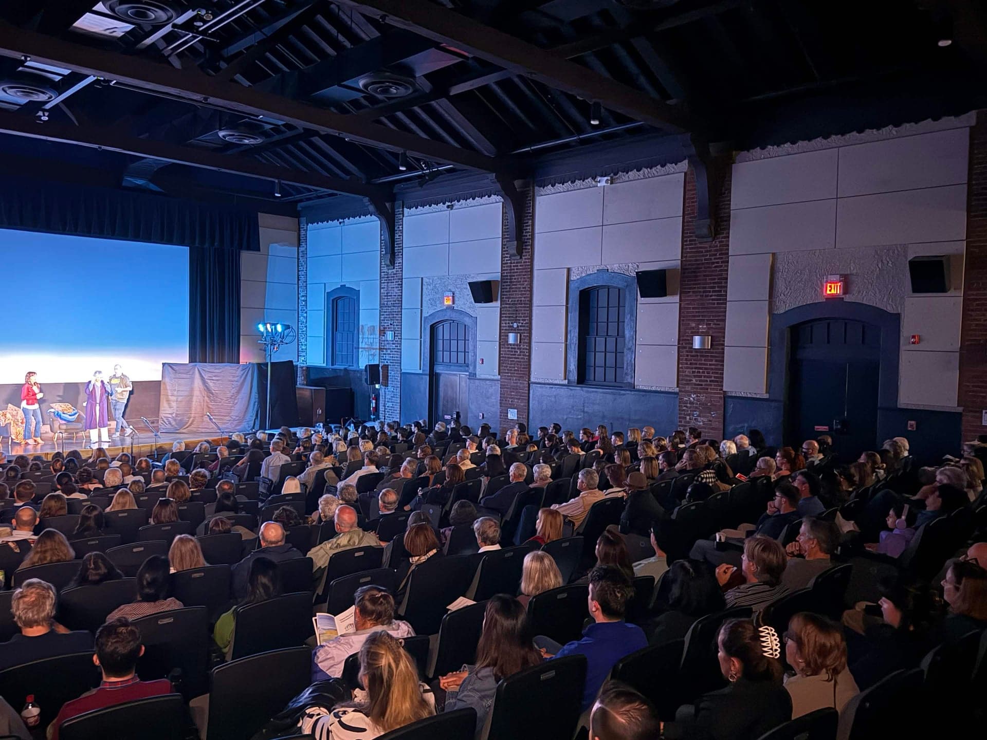 A full house at The Picture House, seen from the side, with three performers on stage at the far left.