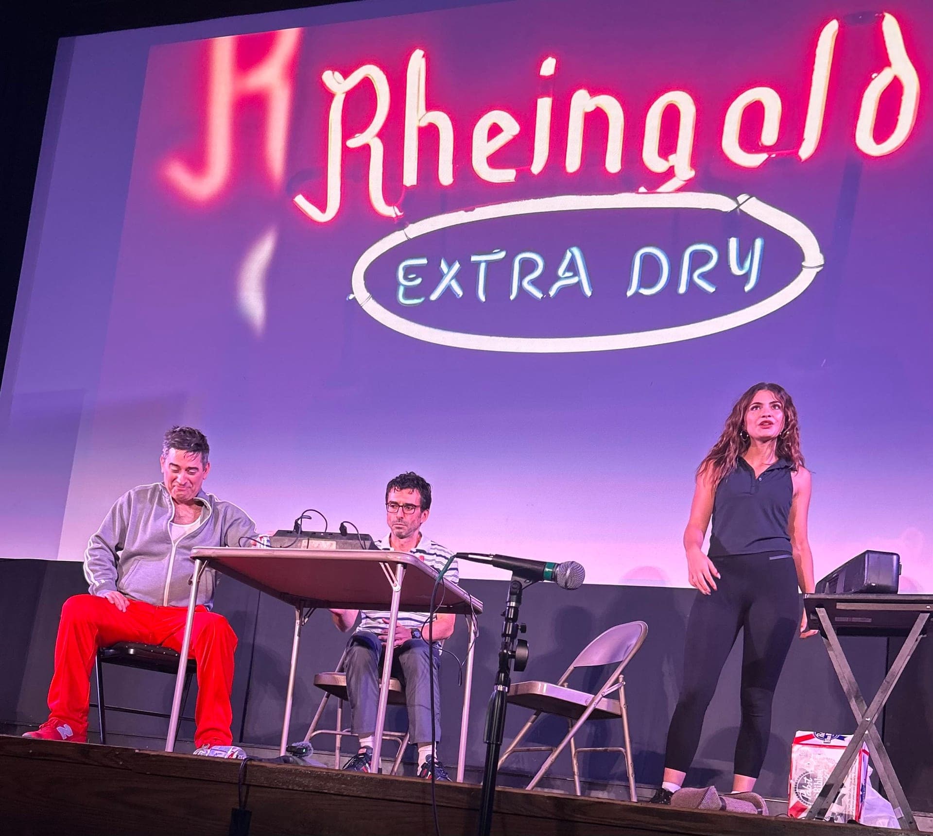 Two men seated at a small table and a woman standing at a microphone, under a projected neon sign reading Rheingold Extra Dry.