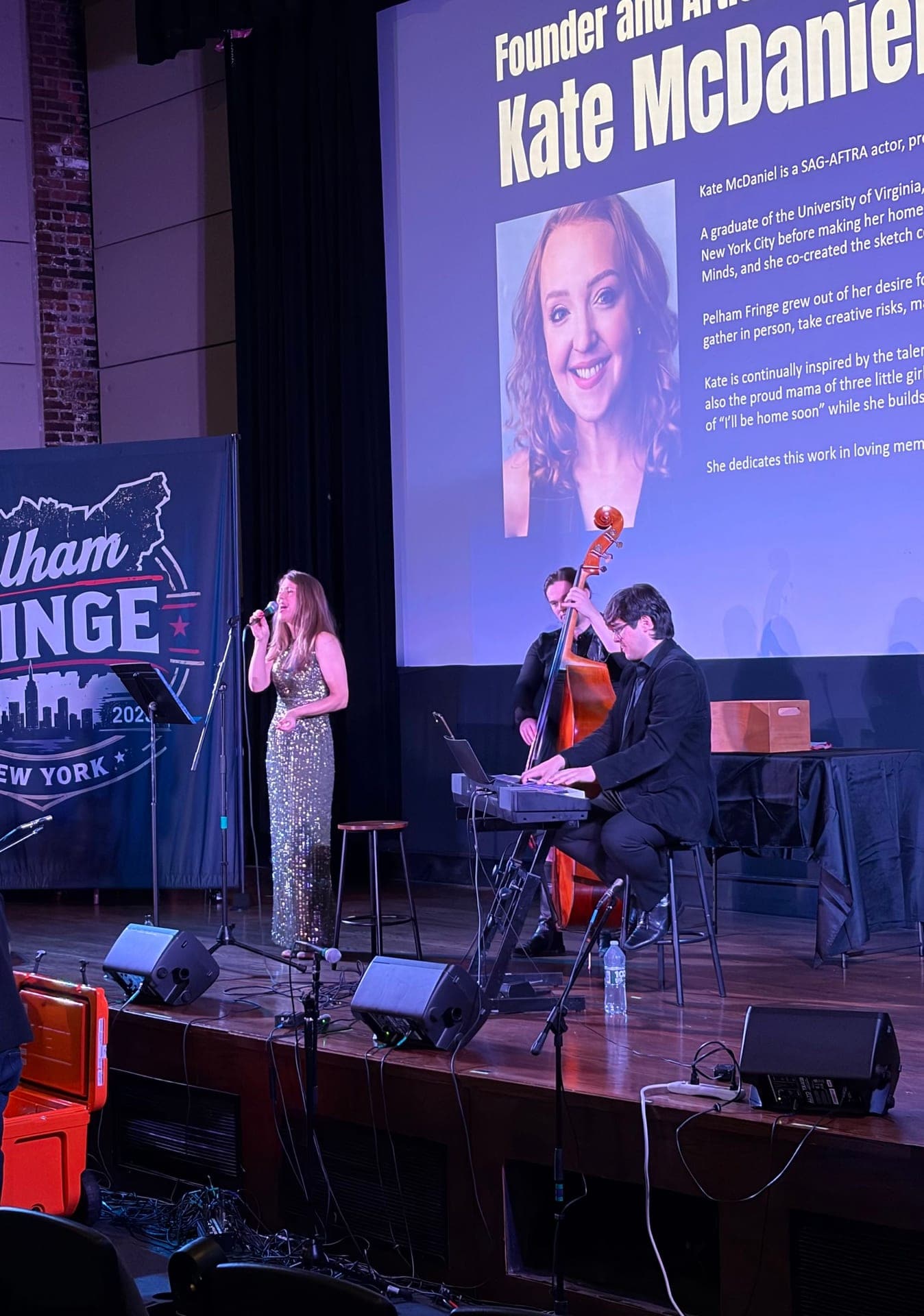 A singer in a sequined jumpsuit at a microphone, with an upright bass player and a keyboard player, and Kate McDaniel's bio projected on the screen above.