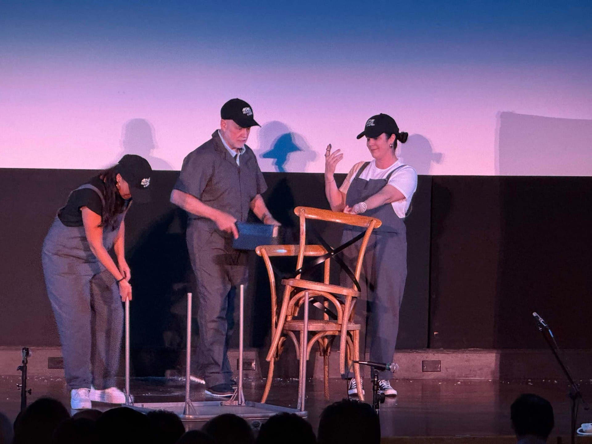 The three ACME improvisers in grey coveralls and black caps moving bistro chairs and a table on stage.