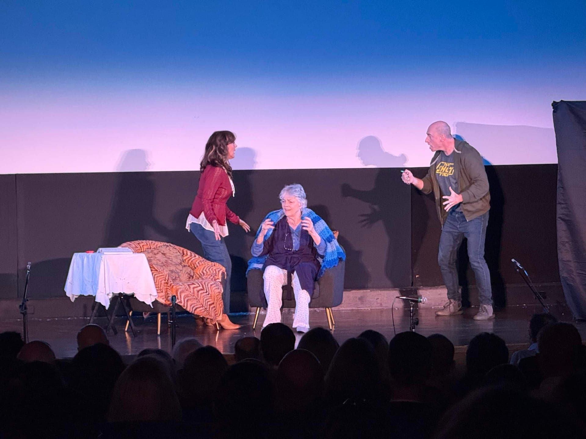 An older woman seated in an armchair under a blue knitted shawl, between a woman in a red cardigan and a man in a green jacket.