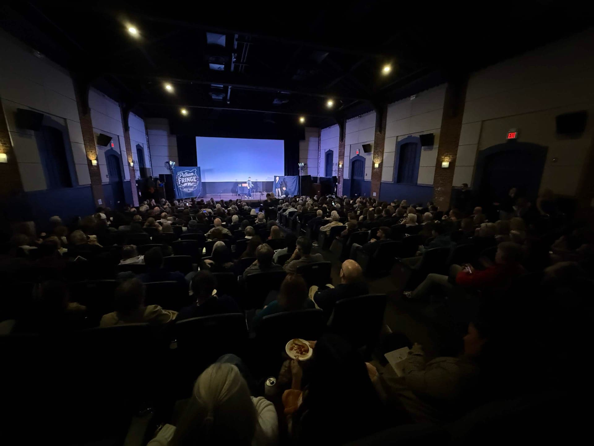 The packed auditorium from the back of the room, rows of audience members facing the lit stage and the Pelham Fringe banner.