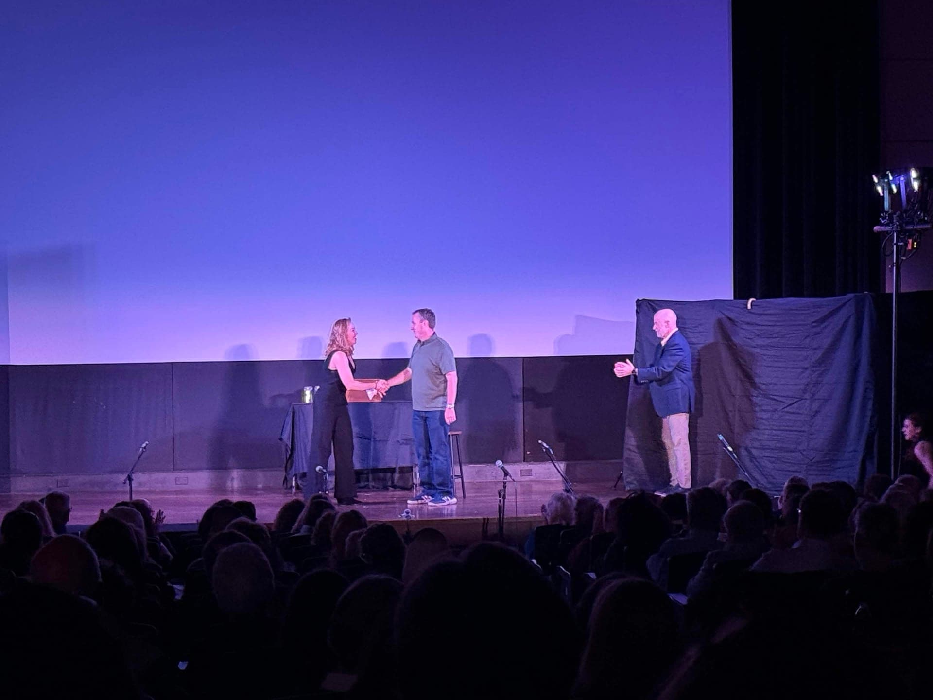 Kate McDaniel shaking hands with Mayor Chance Mullen at center stage, while a man in a blazer applauds at right.