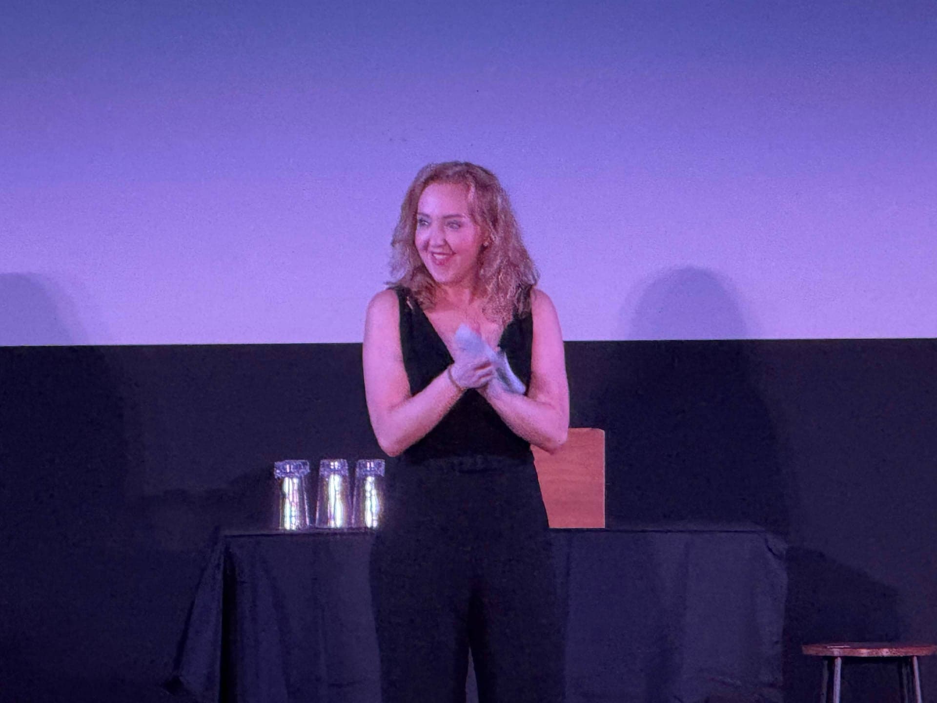 Kate McDaniel smiling on stage in a black jumpsuit, beside a table with three glass jars and a wooden box.