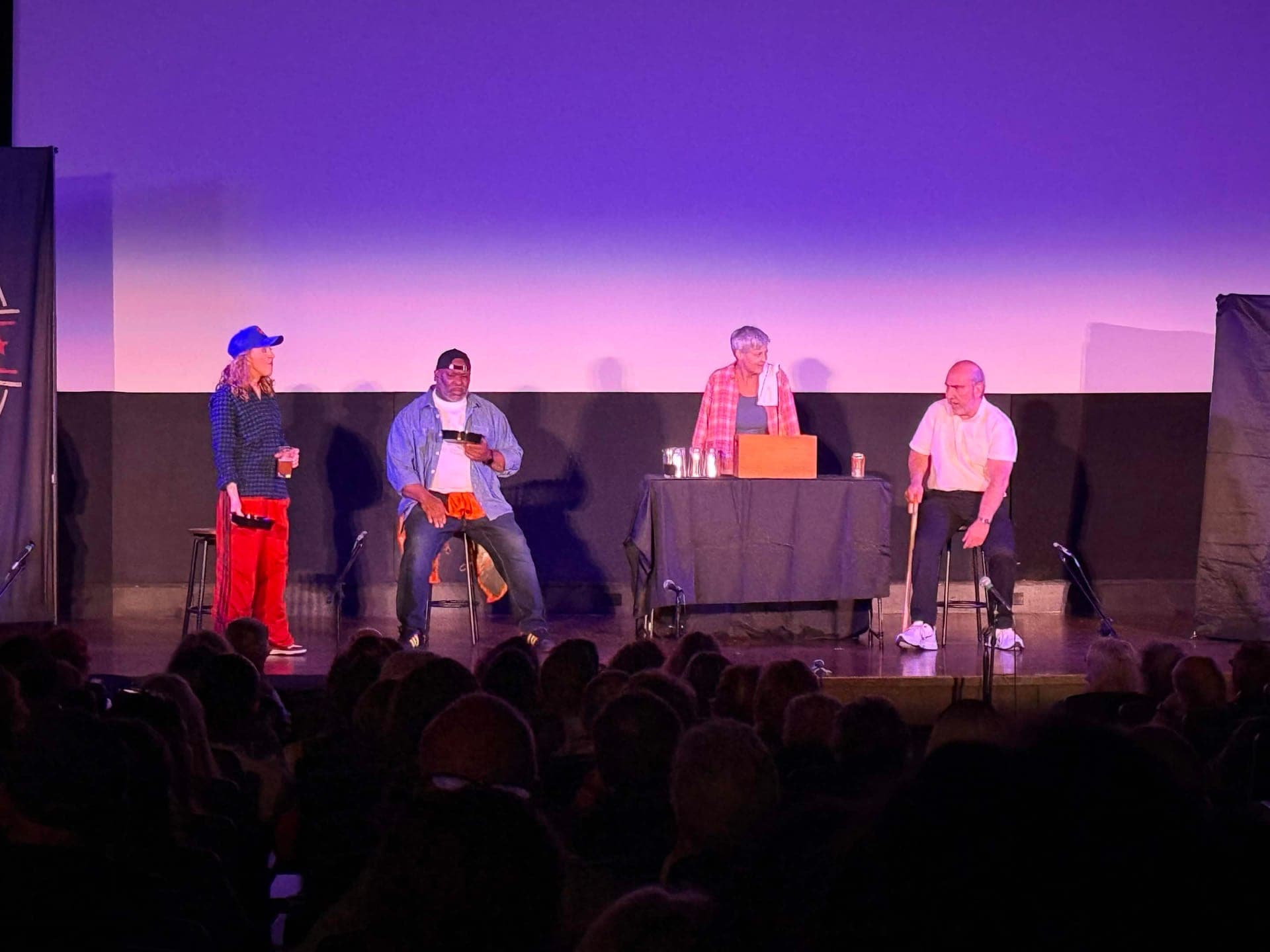Four actors on a purple-lit stage, three seated on stools and one standing behind a draped table with glasses and a wooden box.