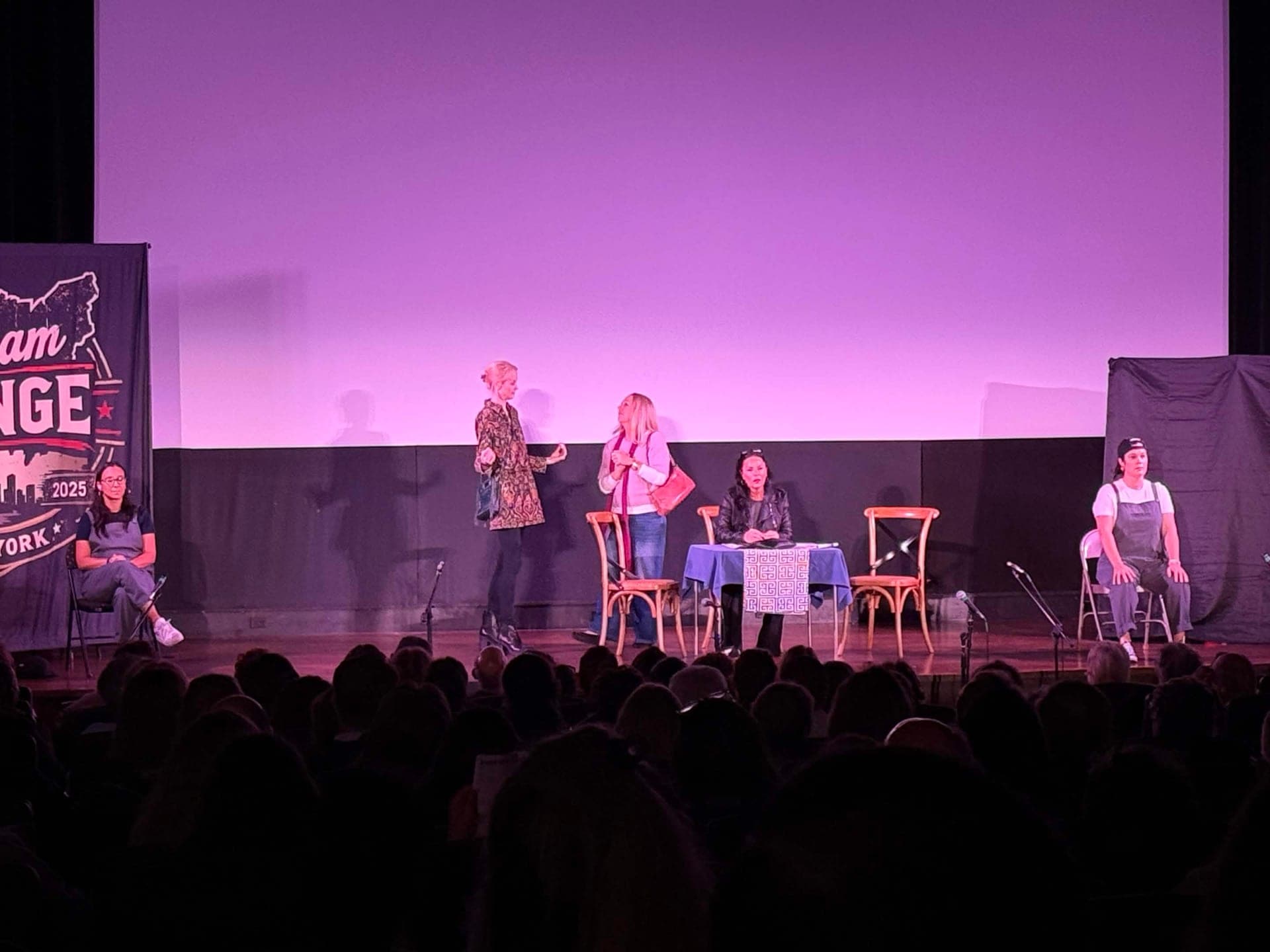 Three actors at a table with a blue cloth in the middle of the stage, with two more cast members in grey coveralls seated at either side.