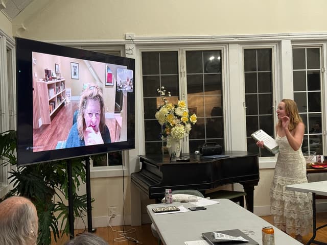 Theresa Rebeck speaking from a video call on a large screen at the front of a room, with a Pelham Fringe member standing to the right holding a script.